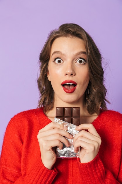 Image of a shocked emotional young woman posing isolated over purple wall wall holding chocolate.