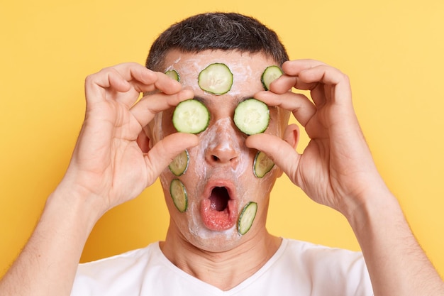 Image of shocked man wearing white t shirt doing cosmetic procedures with cream coveirng eyes with cucumber slices keeps mouth widely opened being astonished isolated over yellow background