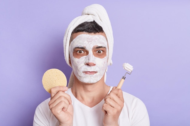 Image of shocked scared man with mask for skin standing with towel on the head holding sponge and massage roller for face isolated over violet background