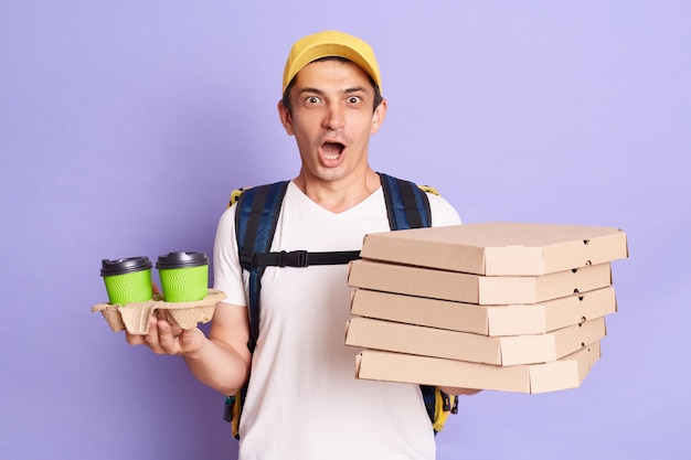 Image of shocked surprised man in yellow cap and tshirt work as dealer courier holding pizza and paper cups of coffee or tea isolated on lilac background