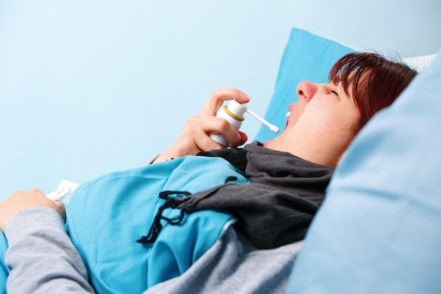 Image of sick brunette using throat spray lying in bed