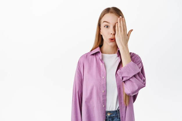 Image of silly beautiful blond girl cover half of face checking vision at optician store pucker lips and look surprised standing against white background