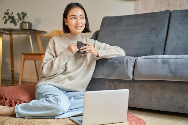 Image of smiling asian woman drinking hot tea holding cup and sitting near laptop on floor resting a