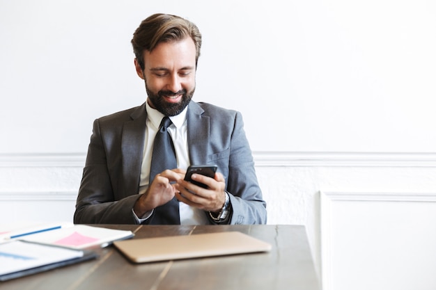 Image of smiling concentrated businessman wearing formal suit typing on cellphone while working in office