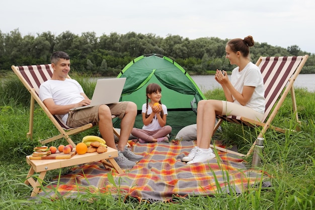 Image of smiling delighted family mother father and their cute daughter being on camping near river man using notebook woman drinking coffee or tea child eating orange