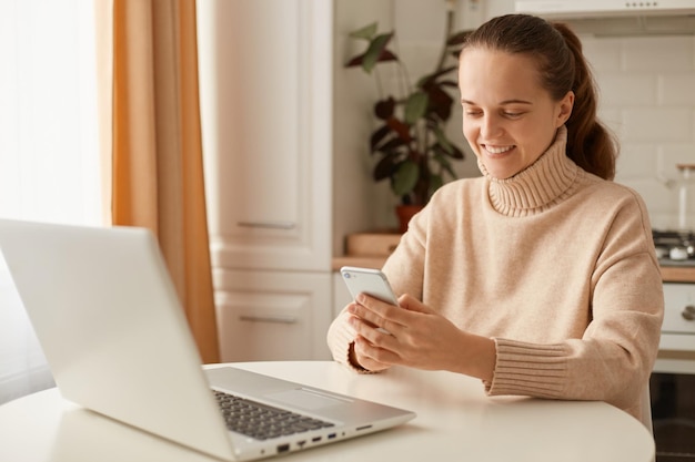 Image of smiling delighted woman wearing beige jumper posing in kitchen female with ponytail holding cell phone in hands looking at mobile phone screen and expressing positive emotions