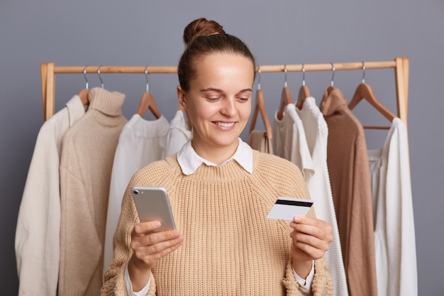 Image of smiling delighted young adult woman in wardrobe standing in front of hanger rack with outfits in mall paying for her shopping online holding mobile phone and credit card in hands