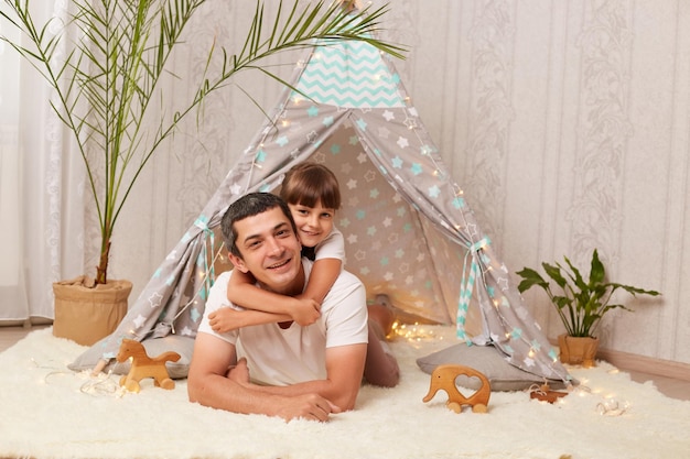 Image of smiling handsome Caucasian man wearing white t shirt lying on floor with his little daughter near her wigwam cute child hugging her father and expressing happiness