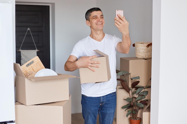 Image of smiling happy man wearing white T-shirt and jeans standing with cardboard box, holding smart phone and taking selfie in a new apartment or broadcasting livestream.