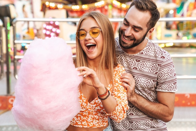 Photo image of a smiling positive loving couple walking outdoors in amusement park eat candyfloss.