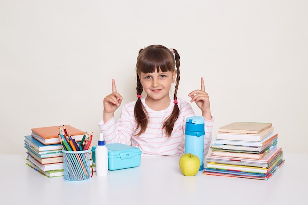 Image of smiling positive optimistic little schoolgirl with dark hair and braids sitting at table surrounded with books pointing fingers up looking at camera posing isolated over white background