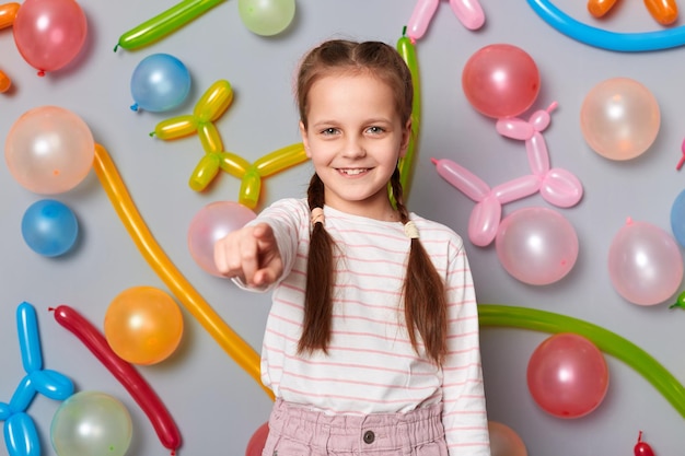 Image of smiling satisfied friendly little girl with braids wearing casual clothing pointing at camera inviting to her party posing isolated over gray background with balloons