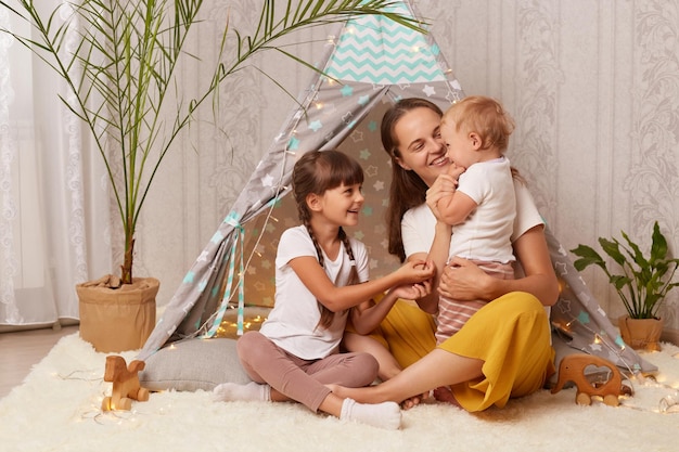 Image of smiling satisfied little girls and her mother playing at home in wigwam and being happy to spend time together female hugging her kids being in good mood