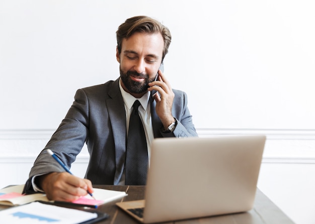 Image of smiling unshaven businessman wearing formal suit talking on cellphone and writing down notes while working at laptop in office