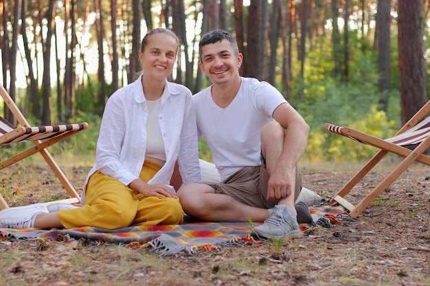 Image of smiling young family enjoying fresh air and beautiful nature in the forest sitting on blanket on the ground looking at camera being happy to spend weekend together