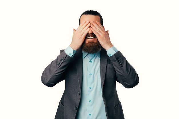 Image of smiling young man in casual suit covering his eyes with palm isolated on white