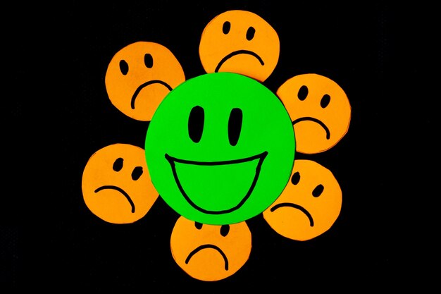 Image of some emoticons cut out of orange paper with a sad face surrounding a big green emoticon with a smiling face concept of bad influences bad friendships negativity problems bullying