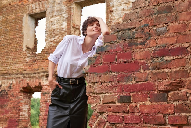 Image of a stylish beautiful woman in a white shirt and leather skirt in a park against the background of a destroyed building The concept of style and fashion