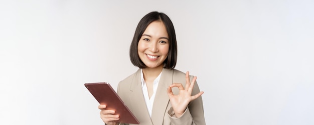 Image of successful asian businesswoman holding digital tablet showing okay ok sign assuring client standing over white background