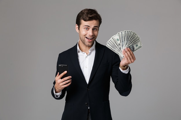 Image of successful businessman 30s in suit holding fan of money and smartphone, isolated over gray wall