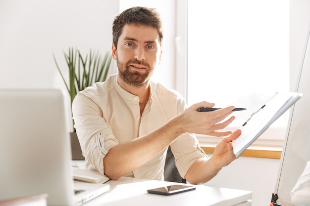 Image of successful office guy 30s wearing white shirt using laptop and paper documents, while working at modern workplace