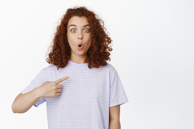 Image of surprised curly woman say wow, look in awe, pointing at herself, standing in tshirt over white background