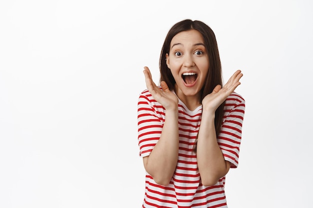 Image of surprised and excited brunette girl, scream from happiness, winning, triumphing and celebrating, standing in t-shirt against white background