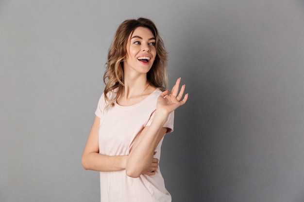 Image of Surprised happy woman in t-shirt waving and looking back over grey