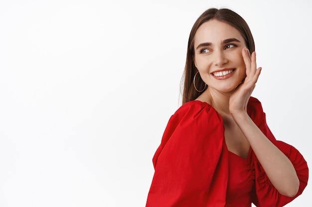 Image of tender and beautiful girl in red romantic dress, wearing evening make up, looking aside at logo promotional text, smiling and gently touch cheek, white background.