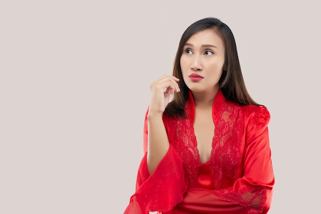 Image of thinking sexy lady standing isolated over pink background. looking away. The hand near the face and mouth. Asian woman in a red satin nightwear wearing lace robe thinking looks left.