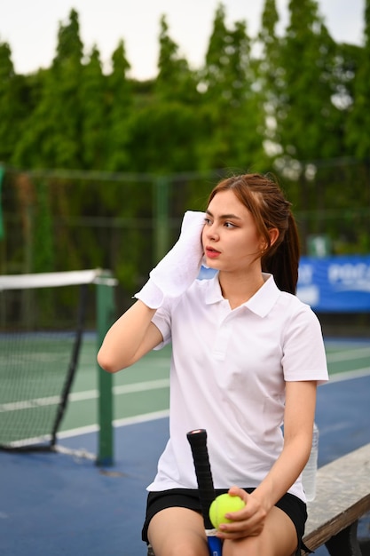 Image of tired female tennis player wiping sweat with towel resting on the bench at the tennis court after training