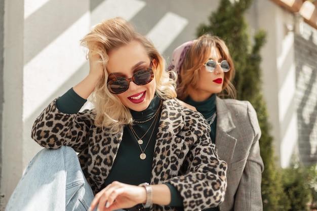 Image of two young happy women friends with fashion sunglasses in trendy vintage urban clothes sits over gray wall with sunlights Looking at camera