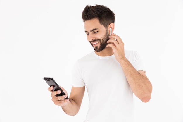 Image of unshaven pleased man listening music with cellphone and earpod isolated over white wall