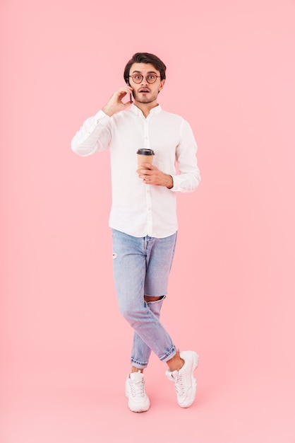 Image of unshaven surprised man wearing eyeglasses drinking coffee takeaway and talking on cellphone isolated