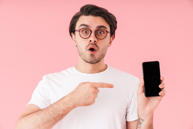 Image of unshaven surprised man wearing eyeglasses holding and pointing finger at cellphone isolated