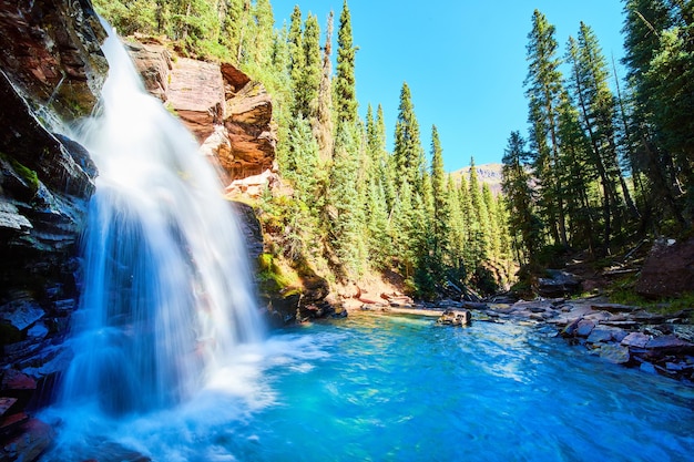 Image of View from side of scenic blue waterfall tucked into gorge in mountains surrounded by pine trees