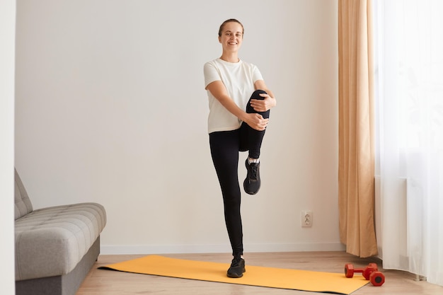 Image of woman exercising by lifting the legs, knees up one by one, holding the knees tightly, standing straight back and hold, relaxing yoga practice at home, looking smiling at camera.