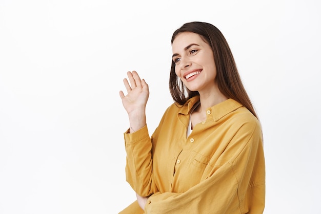 Image of woman standing halfturned waving and looking aside greeting someone on left side copy space say hello hi welcome people standing over white background