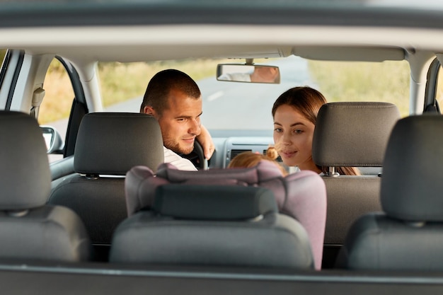 Image of young adult man and woman with little child driving in car baby daughter sitting in car seat father and mother looking back at their child