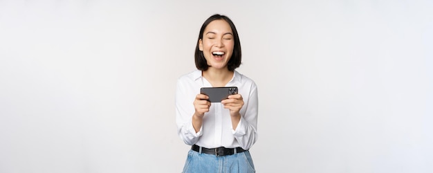 Image of young asian woman watching on smartphone app holding mobile phone and looking at screen laughing and smiling standing over white background