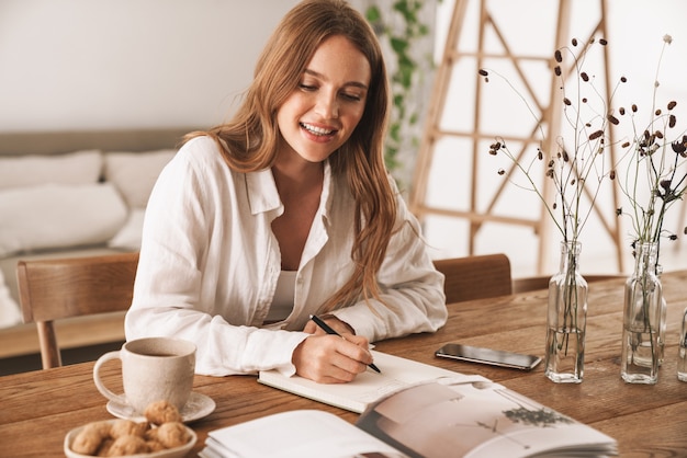 Image of young cheerful happy optimistic woman sit indoors in office writing notes look at magazine.