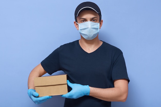 Image of young courier with carton box in hands looking directly at camera isolated over blue wall, online store employee delivers order to client, special service during quarantine.