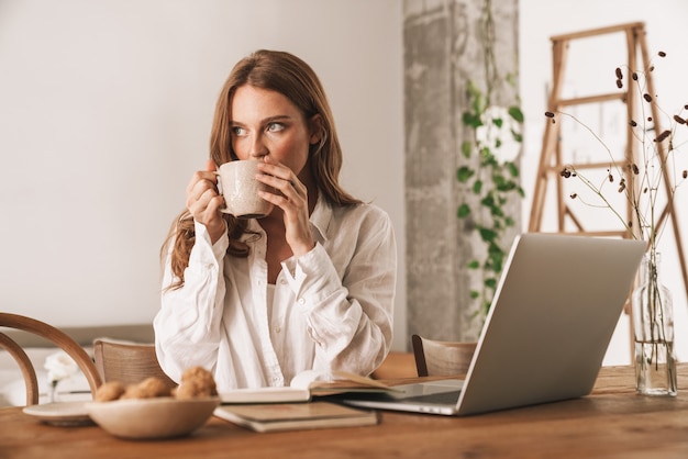 Image of young cute pretty redhead woman sit indoors in office using laptop computer drinking coffee.