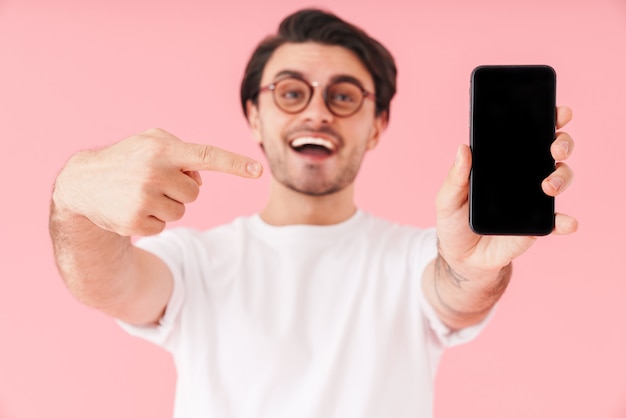 Image of young delighted man wearing eyeglasses holding and pointing finger at cellphone isolated