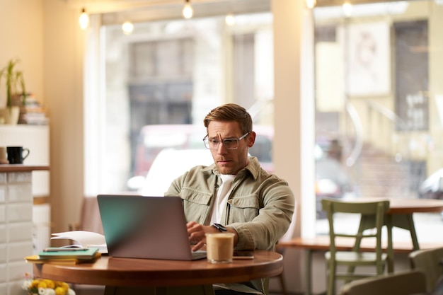 Image of young digital nomad man in glasses sits in cafe works from coffee shop uses laptop in