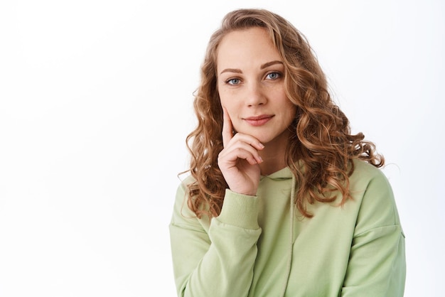 Photo image of young girl student with fair curly hairstyle touching chin and smiling intrigued looking thoughtful thinking about something standing over white background