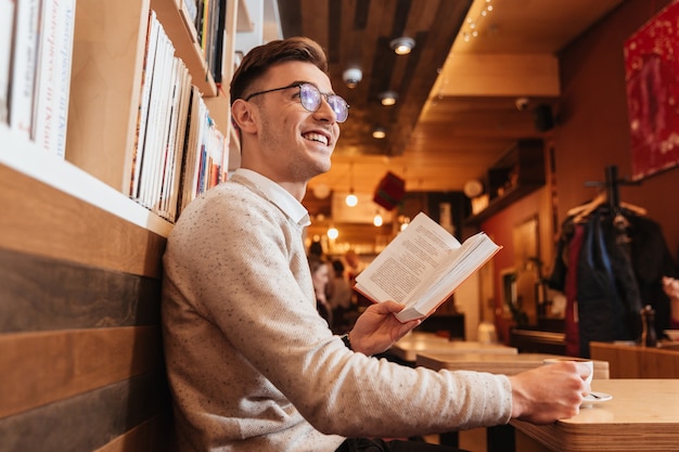 Image of young smiling man sitting in cafe while reading book and look aside.