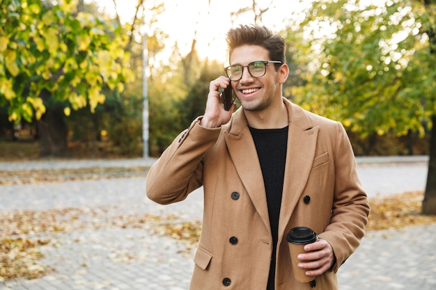 Image of young smiling man wearing coat talking on cellphone and drinking takeaway coffee while walking in autumn park