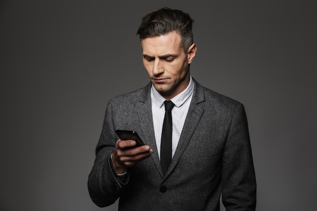 Image of young unshaved man dressed in business costume chatting or working on mobile phone, isolated over gray wall