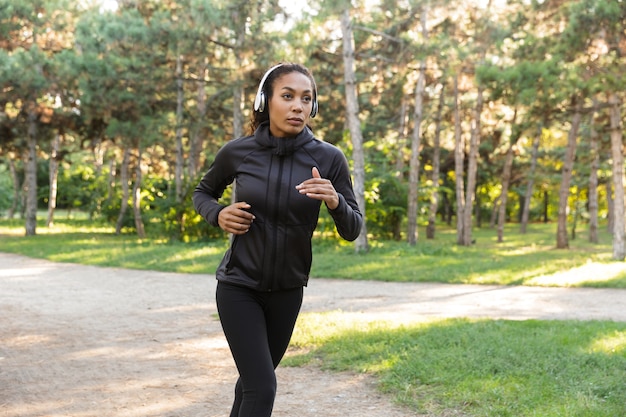 Image of young woman 20s wearing black tracksuit and headphones working out, while running through green park